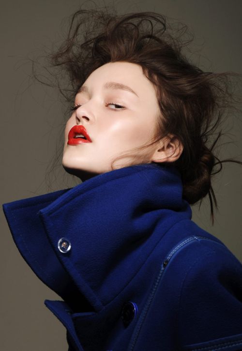 Portrait of a young woman with dark hair and bright red lipstick wearing a dark blue coat against a neutral grey background.