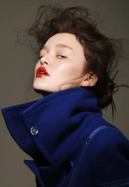 Portrait of a young woman with dark hair and bright red lipstick wearing a dark blue coat against a neutral grey background.