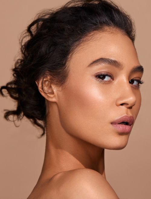 Close-up portrait of a young woman with dark curly hair and neutral makeup against a soft beige background in a beauty or fashion context.
