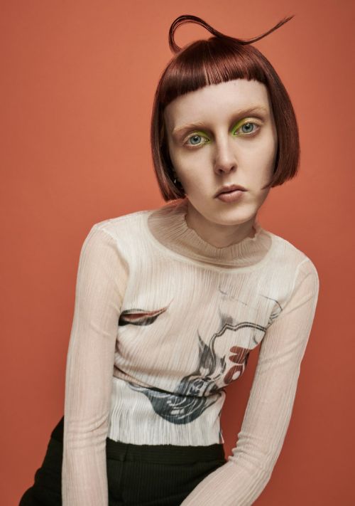 Portrait of a woman with creative hairstyle and makeup, wearing a patterned long-sleeved top against an orange background.