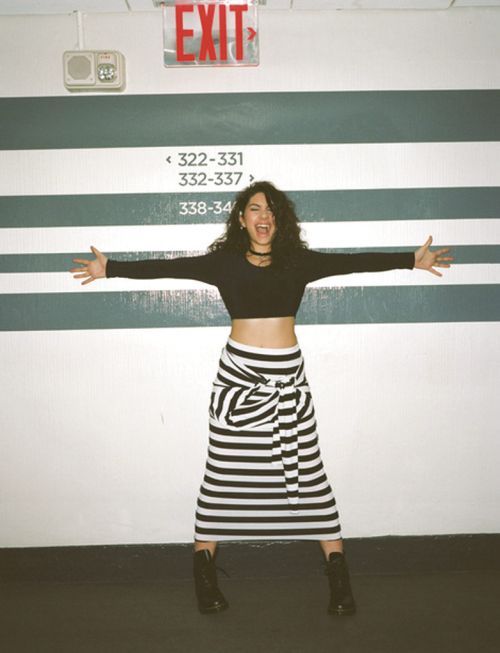 Young woman poses indoors against a striped wall wearing a black crop top, striped skirt, and black boots, smiling with arms outstretched.