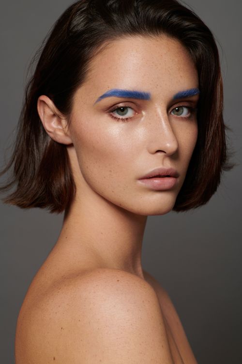Close-up beauty shot of a young adult woman with short brown hair and bright blue painted eyebrows against a neutral gray background.