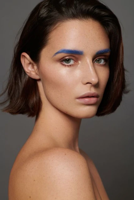 Close-up beauty shot of a young adult woman with short brown hair and bright blue painted eyebrows against a neutral gray background.
