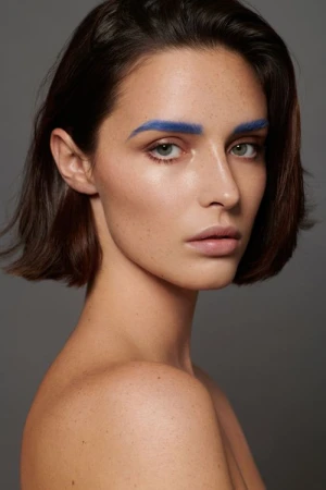 Close-up beauty shot of a young adult woman with short brown hair and bright blue painted eyebrows against a neutral gray background.