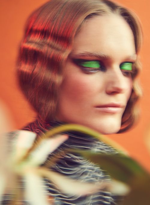 Close-up shot of a woman with green eye makeup and red highlights in her hair, posed in front of an orange backdrop.