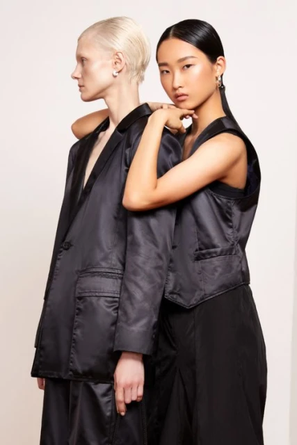 Two women in black satin-like suits pose against a neutral backdrop, one with an arm around the other, sleek hairstyles, and dark suits.