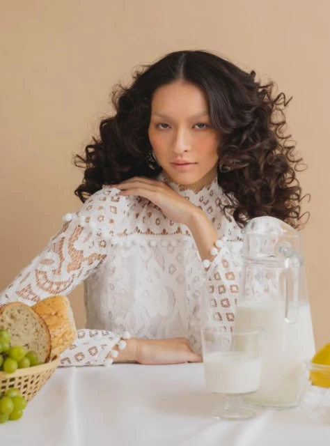 Elegant woman in white lace top sits at a table with a white tablecloth, featuring grapes, bread, milk in glass containers against a neutral background.