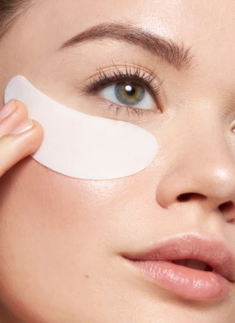Close-up of a woman applying a white under-eye mask. Her face is partially visible, highlighting her eye, cheek, and lips.