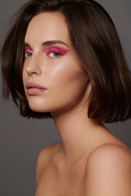 Close-up studio portrait of a young adult female with pink eyeshadow, neutral lipstick, and brown bob haircut against a grey background.