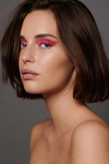 Close-up studio portrait of a young adult female with pink eyeshadow, neutral lipstick, and brown bob haircut against a grey background.
