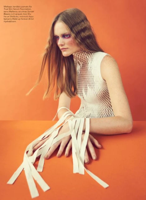 Fashion portrait of a woman with stylized makeup and a net-like top, posed against a vivid orange background. Her hands are wrapped with white ribbons.
