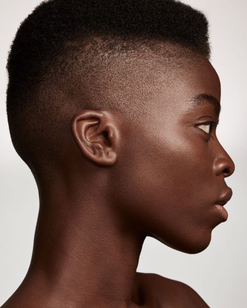 Profile of a young Black person with dark brown skin and short, faded black hair, set against a clean, light grey background.