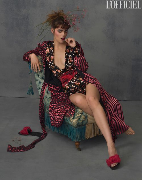 A fashionable woman sits in a vintage chair, wearing a floral robe and fur slippers, with a grey backdrop and editorial lighting.
