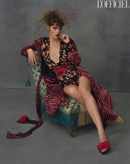 A fashionable woman sits in a vintage chair, wearing a floral robe and fur slippers, with a grey backdrop and editorial lighting.