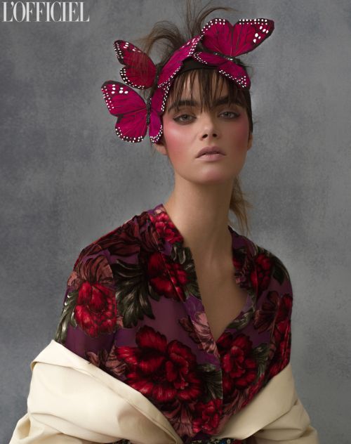 Fashionable woman portrait with magenta butterfly headpiece and floral top against a textured grey background.