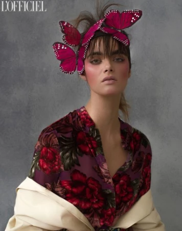 Model wearing large pink butterfly hairpiece and floral blouse with off-white jacket against textured grey background