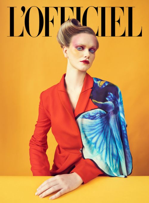 Fashion editorial cover shot: adult woman with stylized eye makeup, red blazer, and blue floral drape in front of a yellow backdrop.