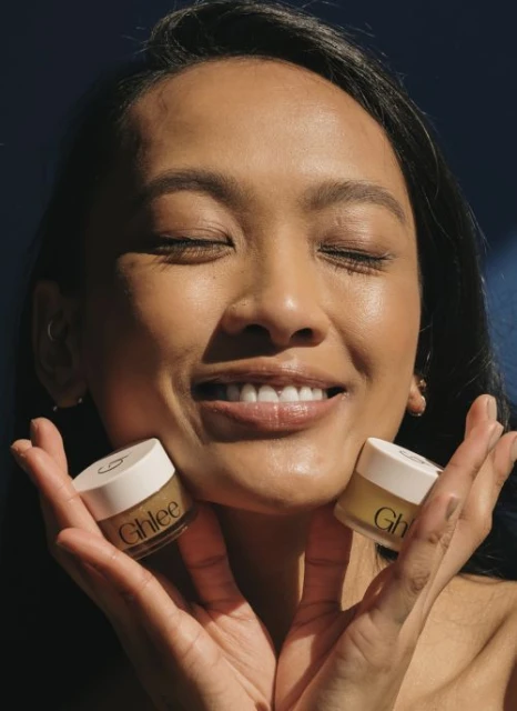 Smiling woman holds two Ghlee skincare jars close to her face, eyes closed, in a beauty product shot with a dark blue background.