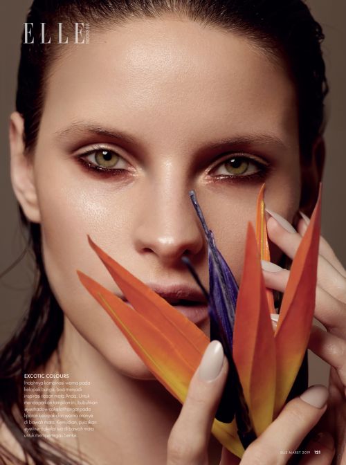 Close-up beauty shot: a poised adult female with vibrant eye makeup and an orange flower against a neutral background, Elle magazine.