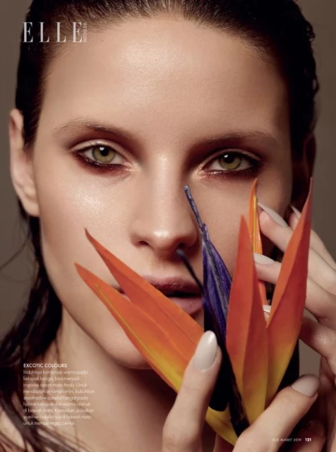 Close-up beauty shot: a poised adult female with vibrant eye makeup and an orange flower against a neutral background, Elle magazine.