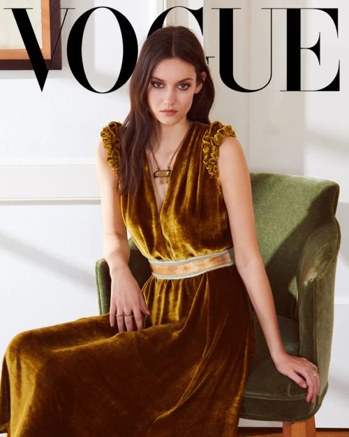 Portrait of a dark-haired woman in a mustard-colored velvet dress on a green chair, as seen on a Vogue magazine cover.