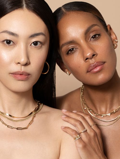 Two beautiful women, one Asian and one Black, are posing indoors while wearing layered gold jewelry against a neutral-toned backdrop.