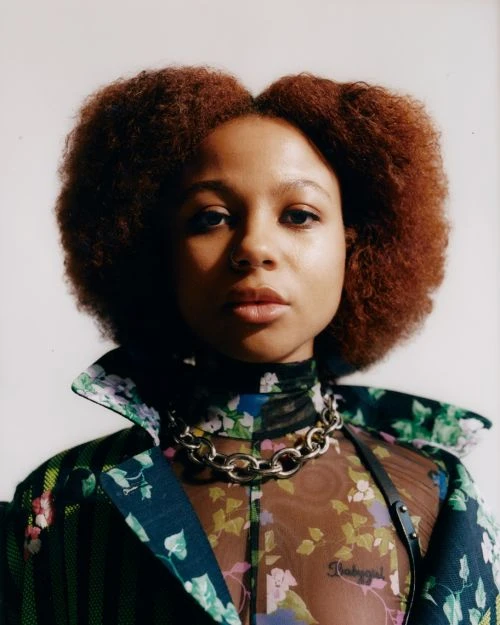 Close-up portrait of a Black woman with auburn hair wearing a floral-print top and a chain necklace against a white background.