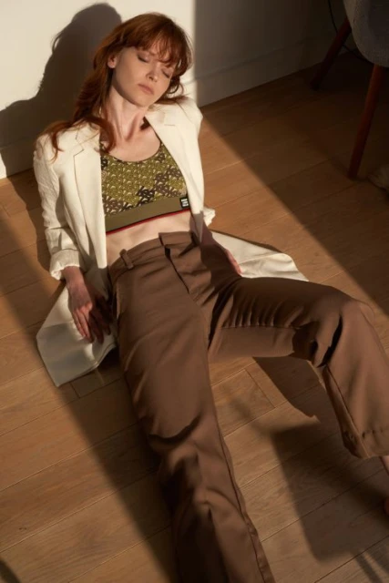A redhead woman wearing a blazer and brown pants is sitting on a wooden floor, eyes closed, illuminated by sunlight streaming through a window.