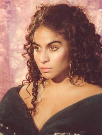 Close-up portrait of a woman with long dark curly hair wearing gold earrings and a dark textured top against a pink iridescent background.