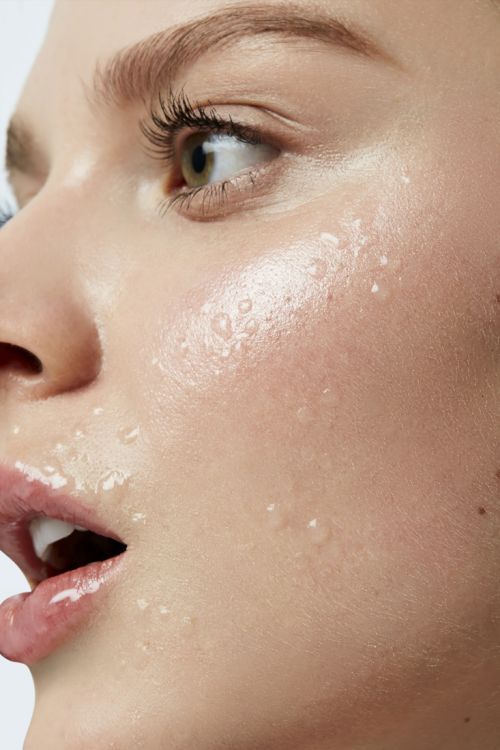 Close-up portrait of a woman's face with water droplets, showcasing her clear skin, green eyes, and parted lips in a beauty context.