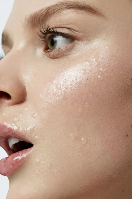Close-up portrait of a woman's face with water droplets, showcasing her clear skin, green eyes, and parted lips in a beauty context.