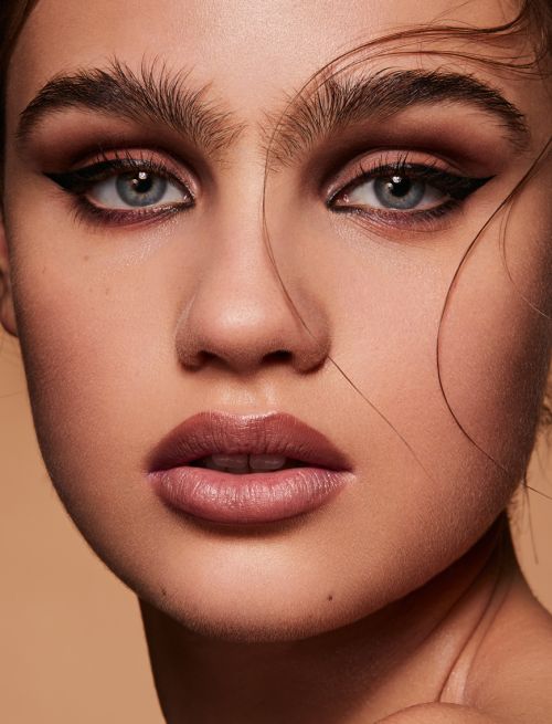 Close-up portrait of a woman with dramatic eye makeup and nude lipstick, showcasing beauty and makeup artistry on a neutral backdrop.