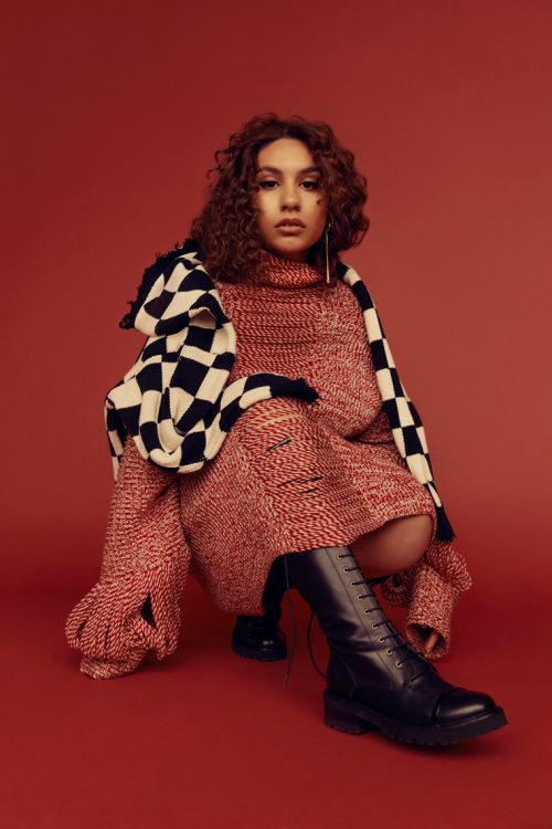 A young woman with curly hair is crouched in a studio setting, wearing a red knit dress, a black and white checkered scarf and boots.