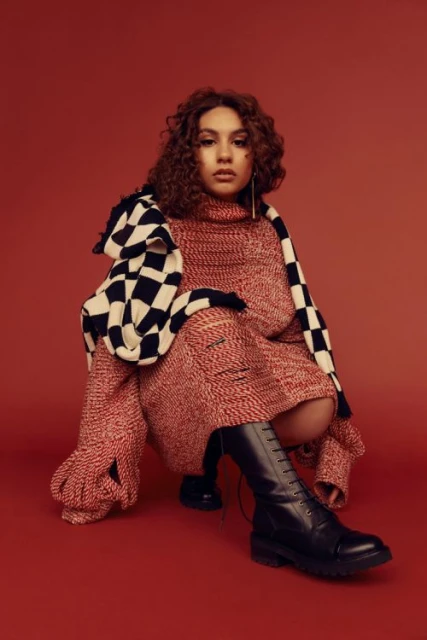 A young woman with curly hair is crouched in a studio setting, wearing a red knit dress, a black and white checkered scarf and boots.