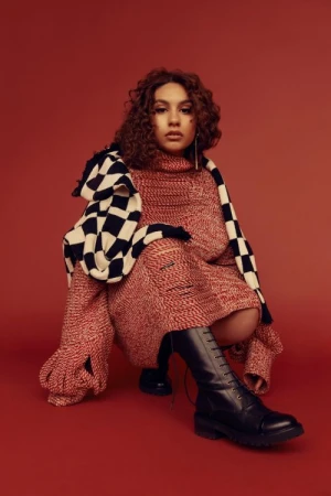 A young woman with curly hair is crouched in a studio setting, wearing a red knit dress, a black and white checkered scarf and boots.