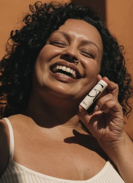 Close-up of a smiling adult female holding a tube of white lip balm against a solid tan background in natural light.