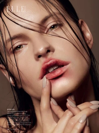 Close-up portrait of a woman with wet hair and blurred red lipstick, touching her chin with long nude fingernails.