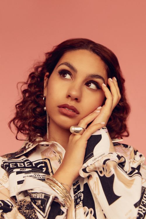 Attractive young woman with brown curly hair looks upward in a studio shot with a pink backdrop while wearing a graphic shirt.