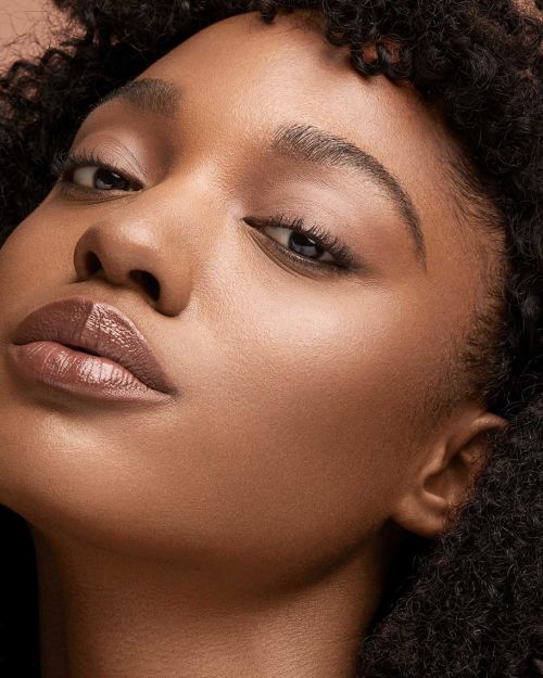 Close-up portrait of a young Black woman with dark curly hair, warm skin, and light pink lipstick, posing against a dark background.