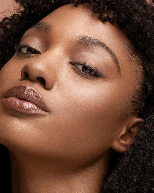 Close-up portrait of a young Black woman with dark curly hair, warm skin, and light pink lipstick, posing against a dark background.