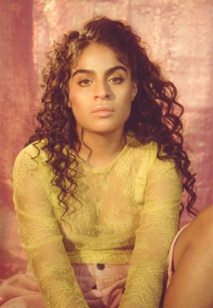 Portrait of a woman with long curly brown hair wearing a sheer yellow top against a pink background