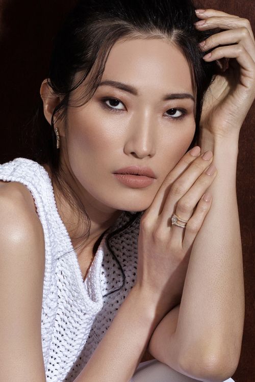 Close-up portrait of an Asian adult female model with dark hair, wearing a white knit top and rings, posing in a studio with brown background.