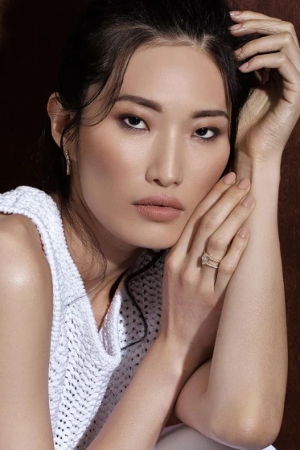 Close-up portrait of an Asian adult female model with dark hair, wearing a white knit top and rings, posing in a studio with brown background.