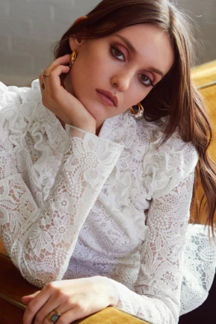 Portrait of a brunette adult woman in a white lace blouse with long sleeves. She is wearing gold hoop earrings and rings on her fingers.