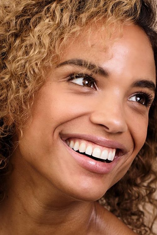 Close-up of a smiling adult female with curly hair, showcasing a joyful and engaging expression in a well-lit, natural setting.