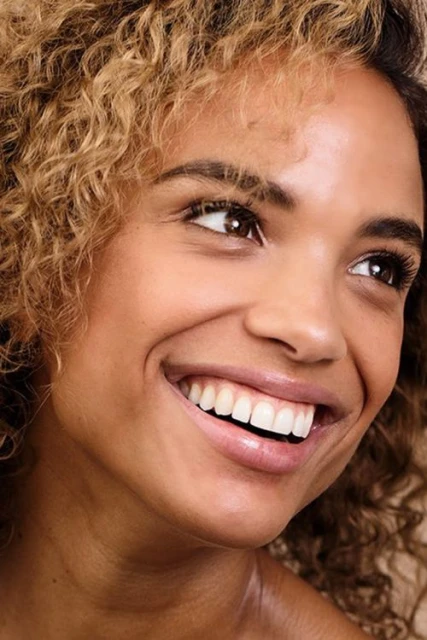 Close-up of a smiling adult female with curly hair, showcasing a joyful and engaging expression in a well-lit, natural setting.