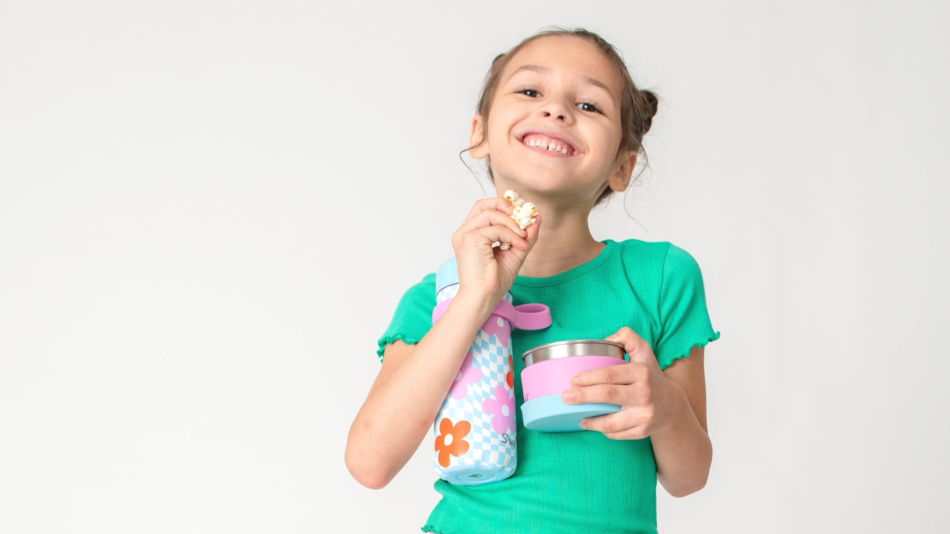 Smiling child in a green shirt eats popcorn and poses in a studio with a lunch container and water bottle on white backdrop.