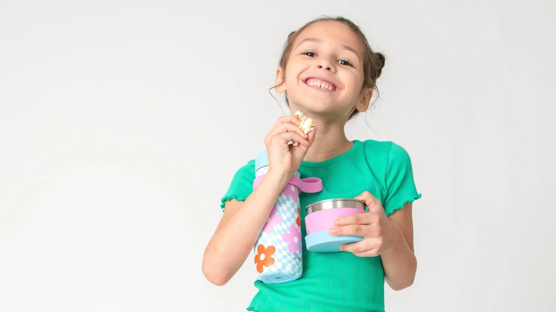 Smiling child in a green shirt eats popcorn and poses in a studio with a lunch container and water bottle on white backdrop.