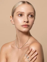 Close-up portrait of a woman with sleek blonde hair, wearing multiple silver earrings, rings, and a beaded necklace against a beige background.