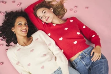 Two smiling women lying down head to head on a pink background wearing heart-patterned sweaters and jeans.